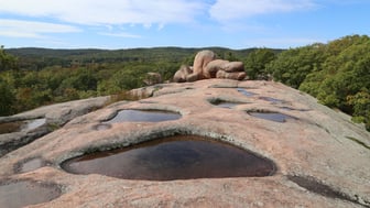 Weathered pink granite dome at Elephant Rocks State Park, Missouri, with rainwater pooled in shallow basins and a cluster of giant rounded boulders beyond, above forested hills under a bright sky.