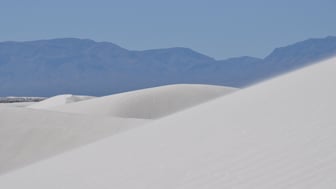 Rippled white gypsum dunes at White Sands National Park, New Mexico, with a hazy blue mountain range in the distance under a clear sky.