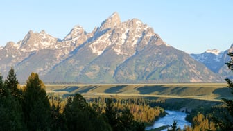 The snow-streaked granite peaks of the Teton Range rise above forested slopes and open sagebrush flats, with a bend of the Snake River winding through cottonwoods in the foreground under a clear morning sky.